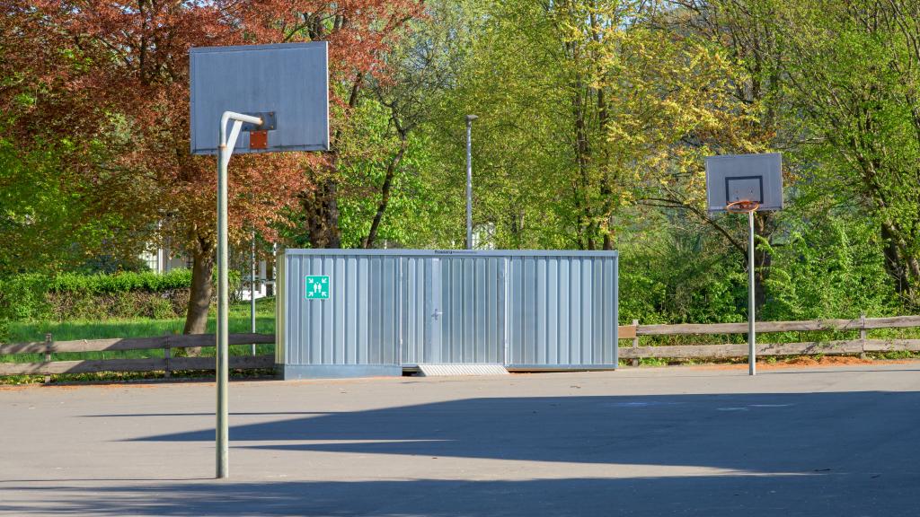 Gesamtansicht des Schulhofs der Grundschule Dattenfeld mit Basketballplatz und dem neuen Spielzeug- und Materialcontainer, der sich harmonisch in das Umfeld einfügt.