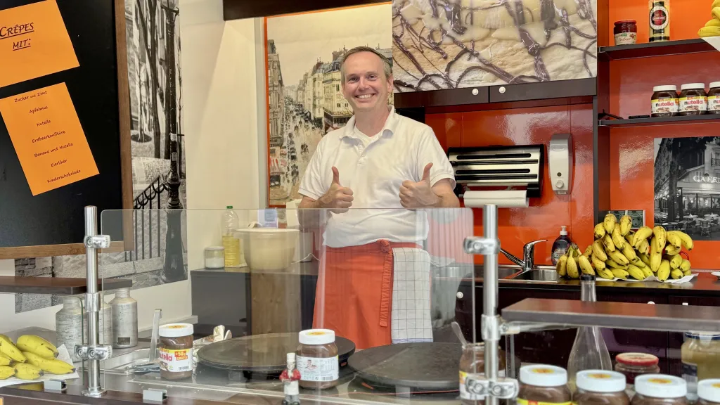 Ein lächelnder Crêpe-Bäcker steht mit zwei erhobenen Daumen hinter seinem Verkaufsstand mit Crêpe-Platten, Nutella und Bananen.